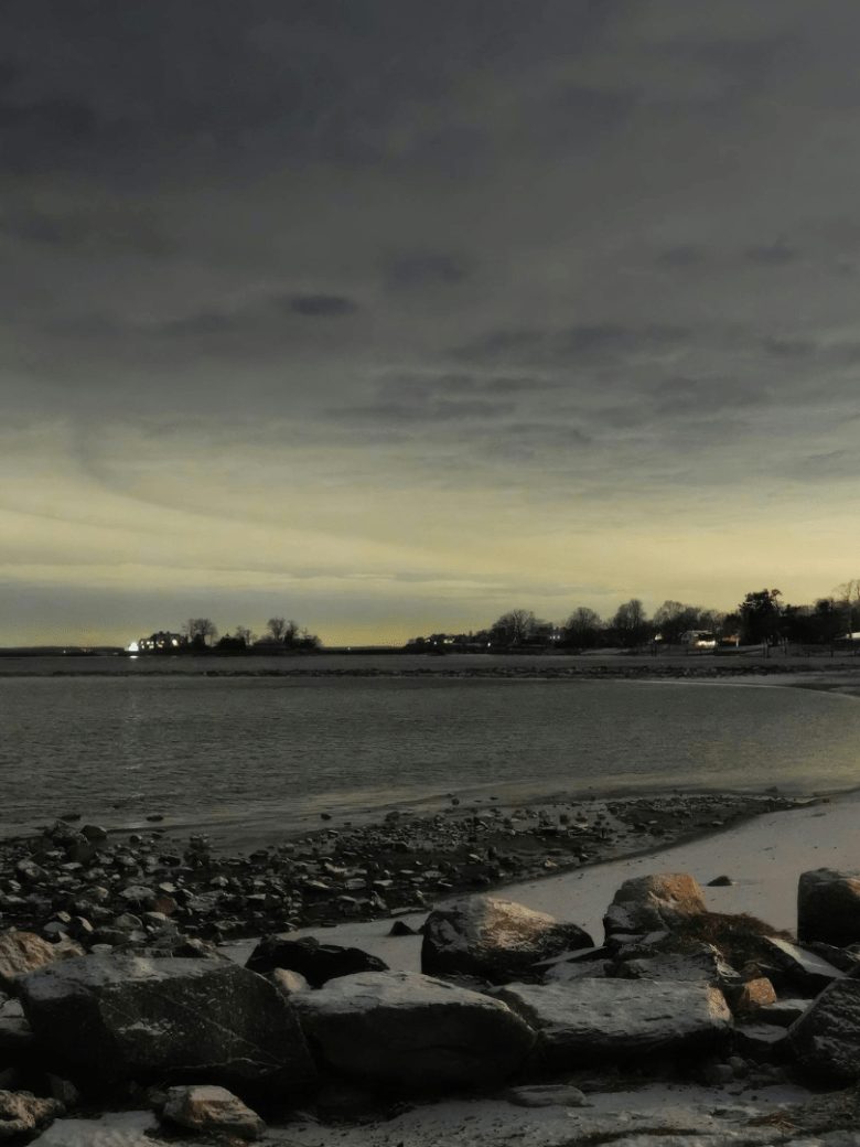 Night view of a rocky beach under a cloudy sky, with distant houses lit on the horizon.